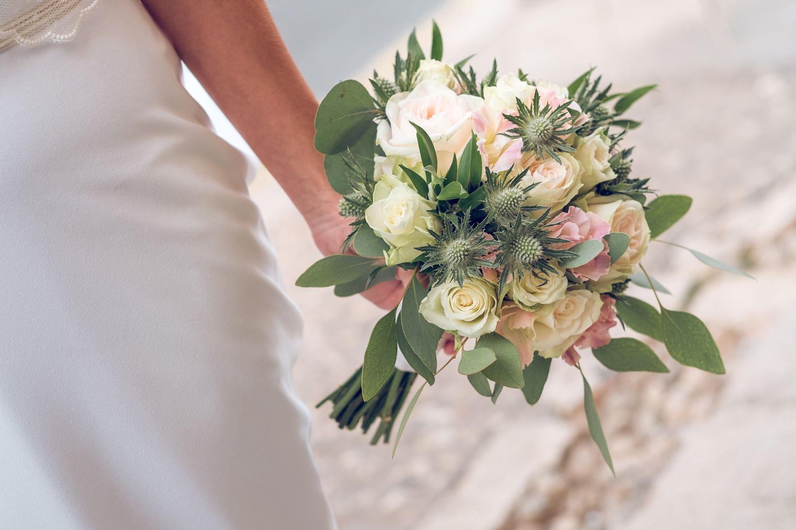 A bride in a white gown carrying a bouquet of cream roses, thistle and greenery at her side