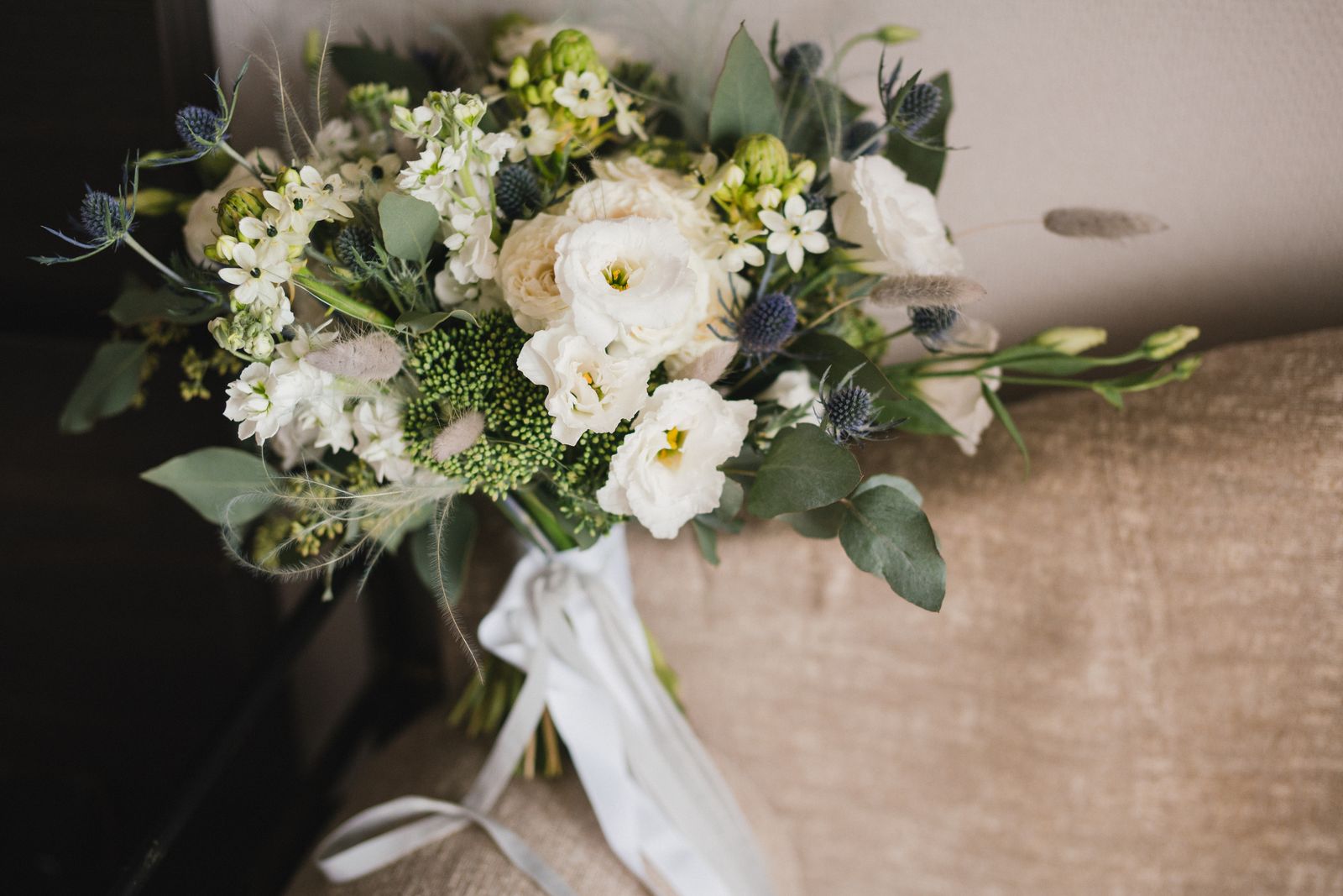 A bridal bouquet of white lisianthus, roses and blue thistle tied with a white silk ribbon